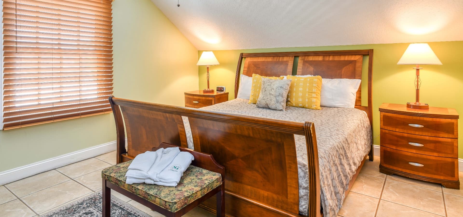 A cozy bedroom with a wooden bed, decorative pillows, bedside lamps, and a bench with towels.