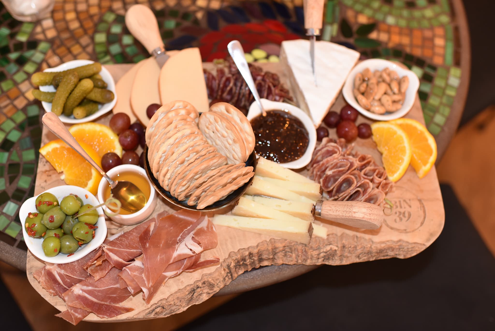 A wooden platter displays an assortment of cheeses, cured meats, olives, pickles, fruit, and spreads.