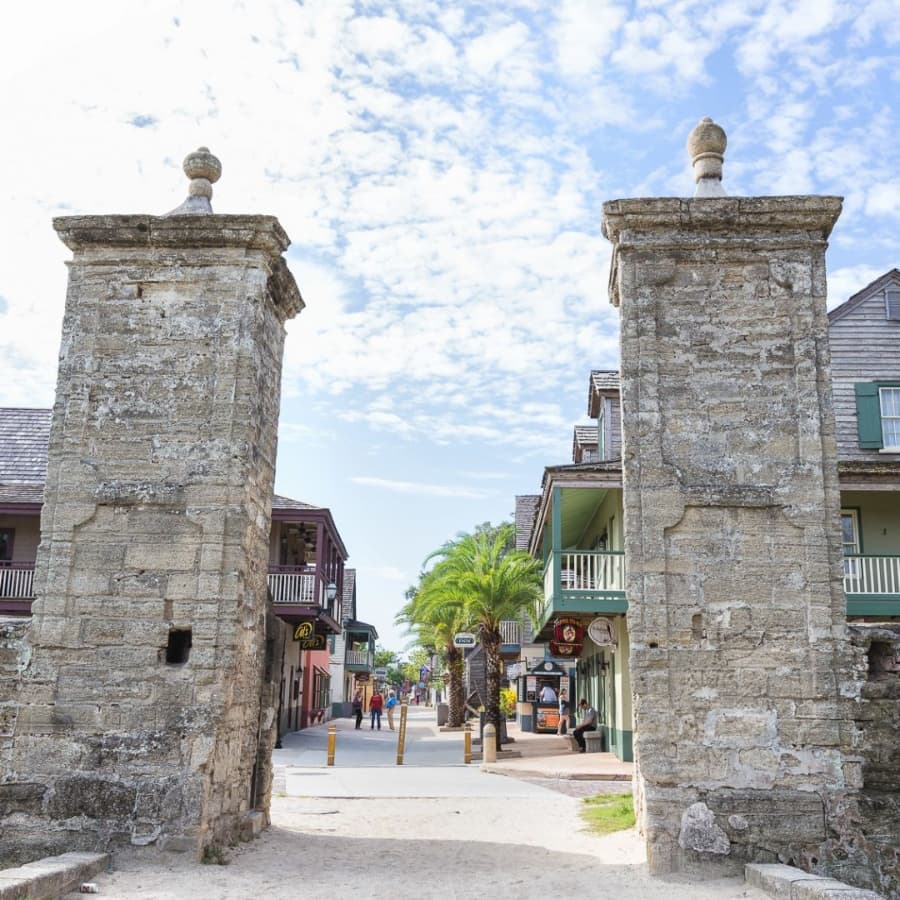 Historic stone gate leading into a vibrant street lined with shops and palm trees.