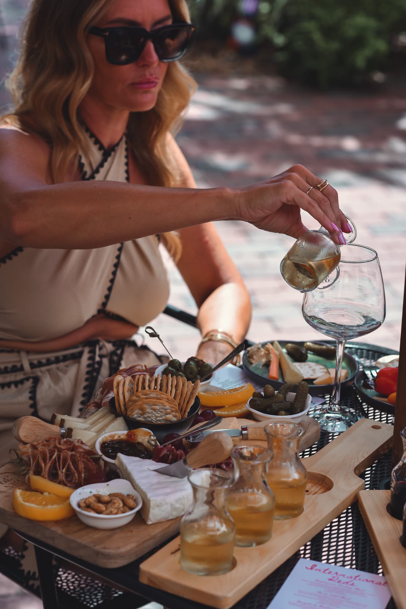 A woman in sunglasses pours a drink into a glass while surrounded by a charcuterie board and small glass bottles.