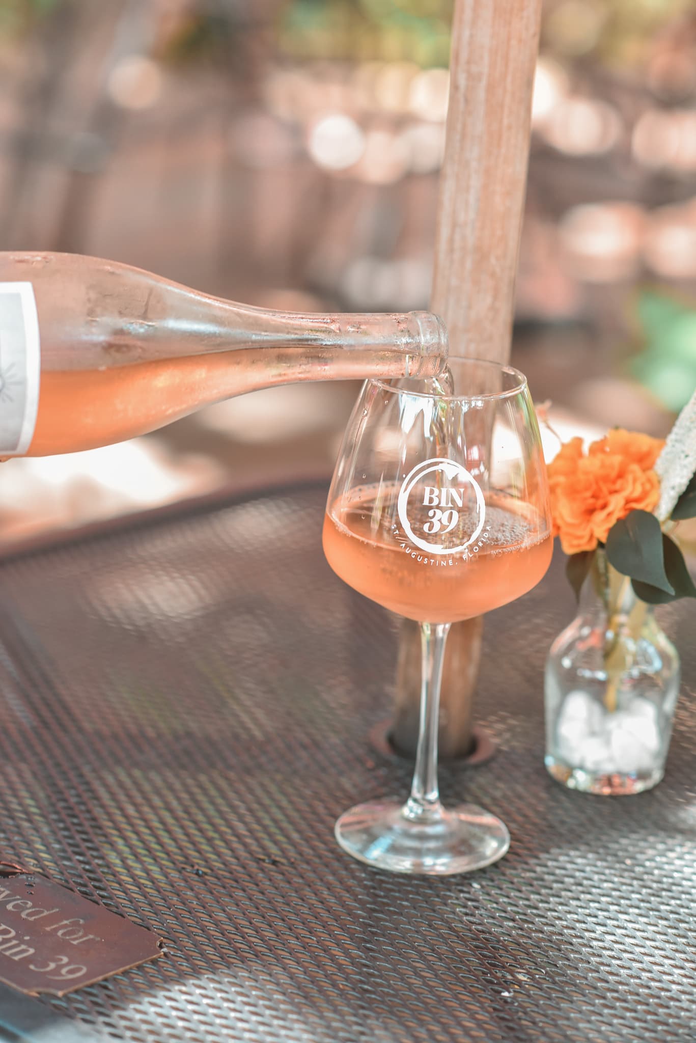 A bottle pours a pink drink into a branded glass on a table with a flower vase.