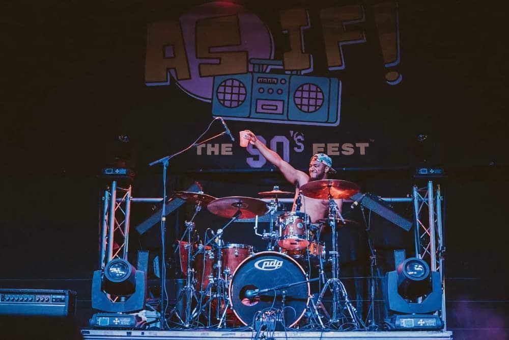 A drummer performs energetically on stage at a music festival backdrop.