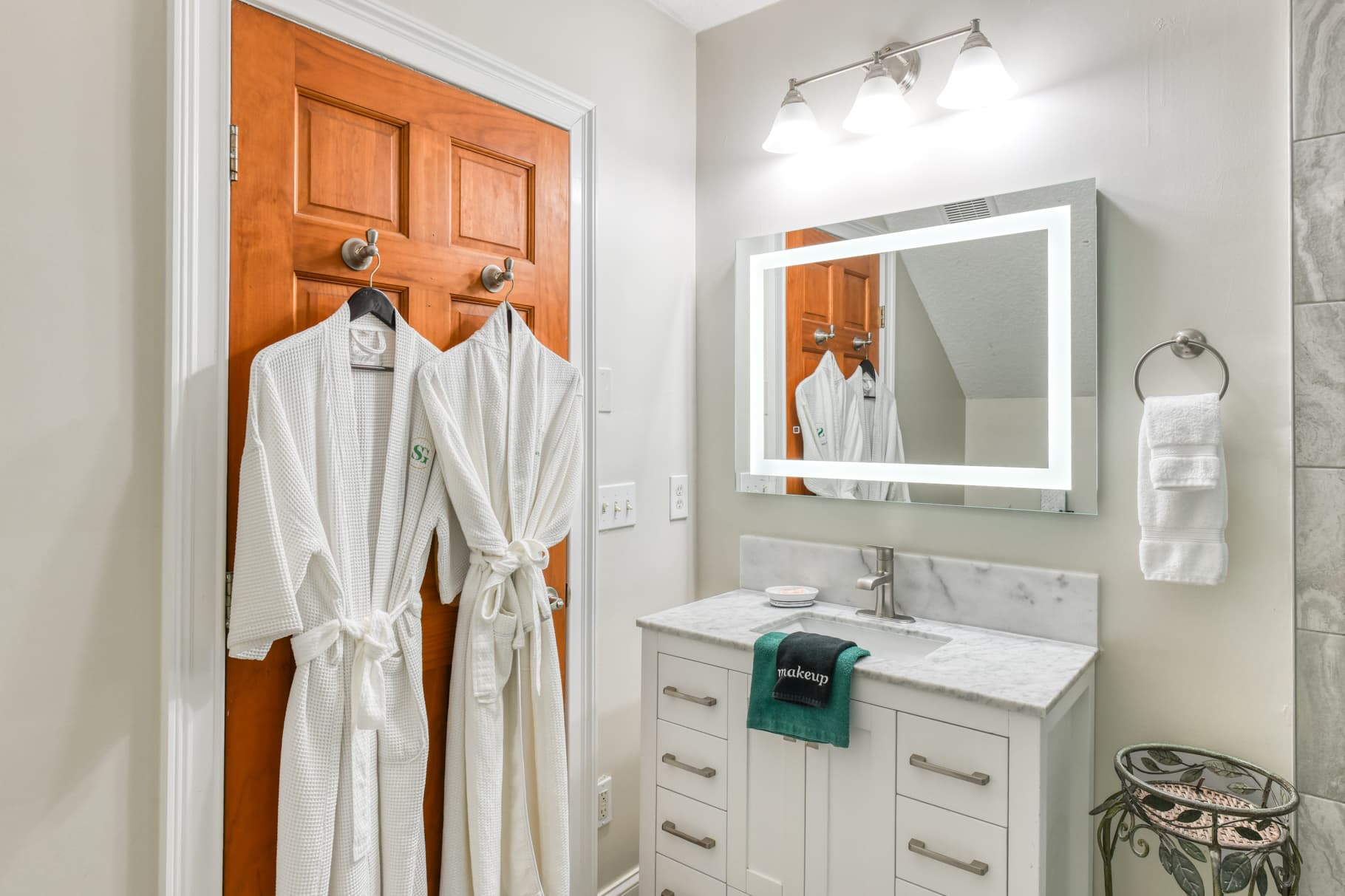A bright bathroom featuring two hanging white robes, a sleek vanity with a marble countertop, and a mirror with integrated lighting.