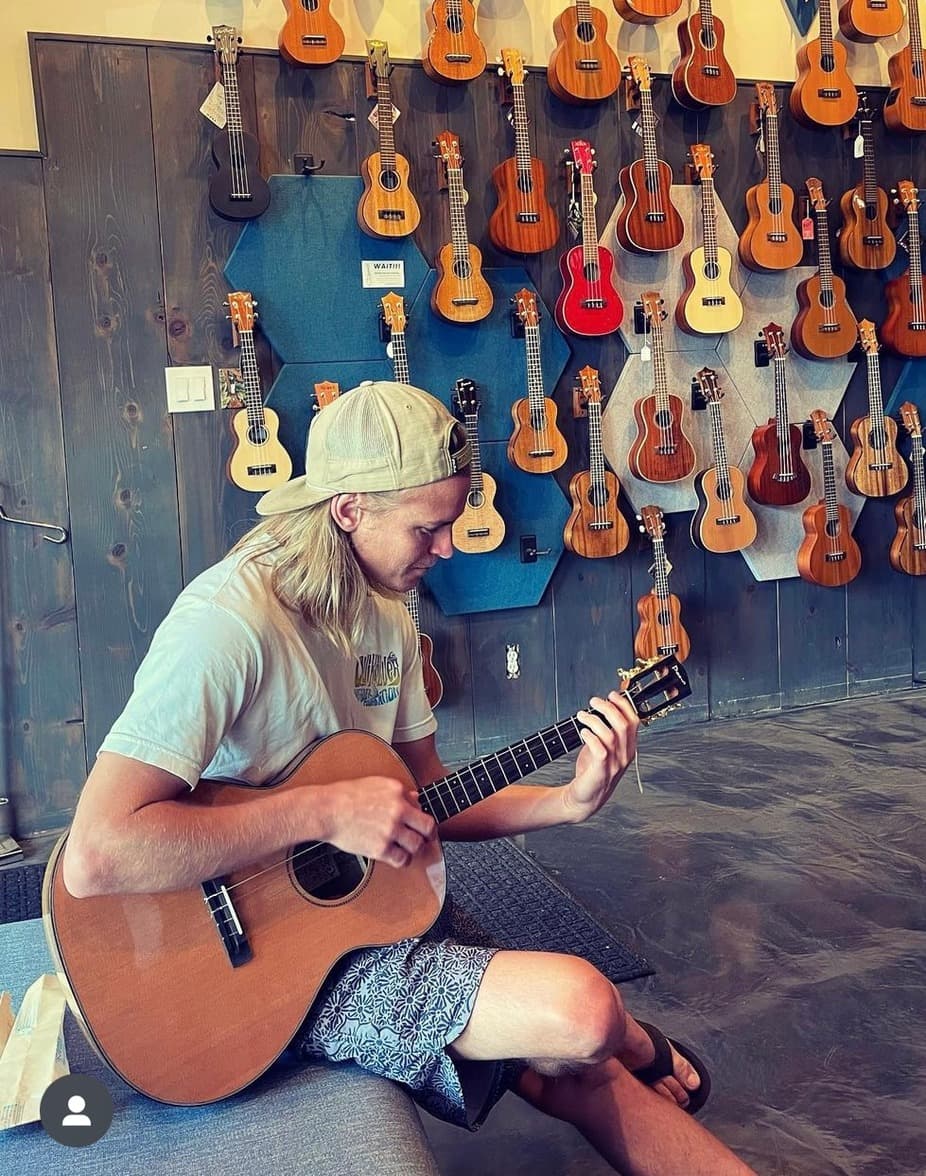 A young man with long hair plays a guitar in a shop filled with hanging ukuleles.