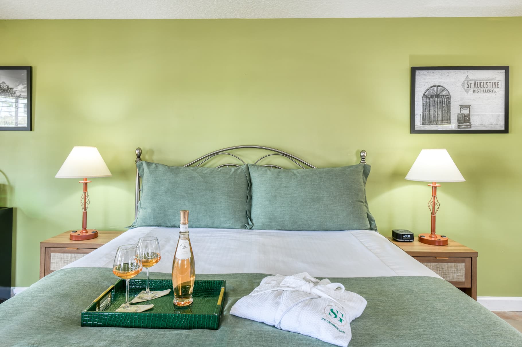 A cozy bedroom featuring a green accent wall, a neatly made bed with pillows, a tray of wine and glasses, and warm lighting.