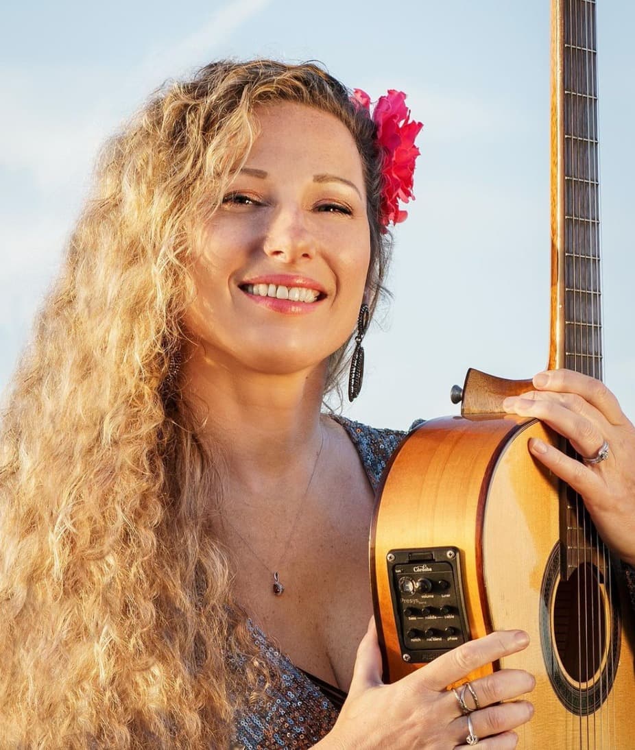 A smiling woman with long curly hair holds an acoustic guitar, adorned with a flower in her hair.