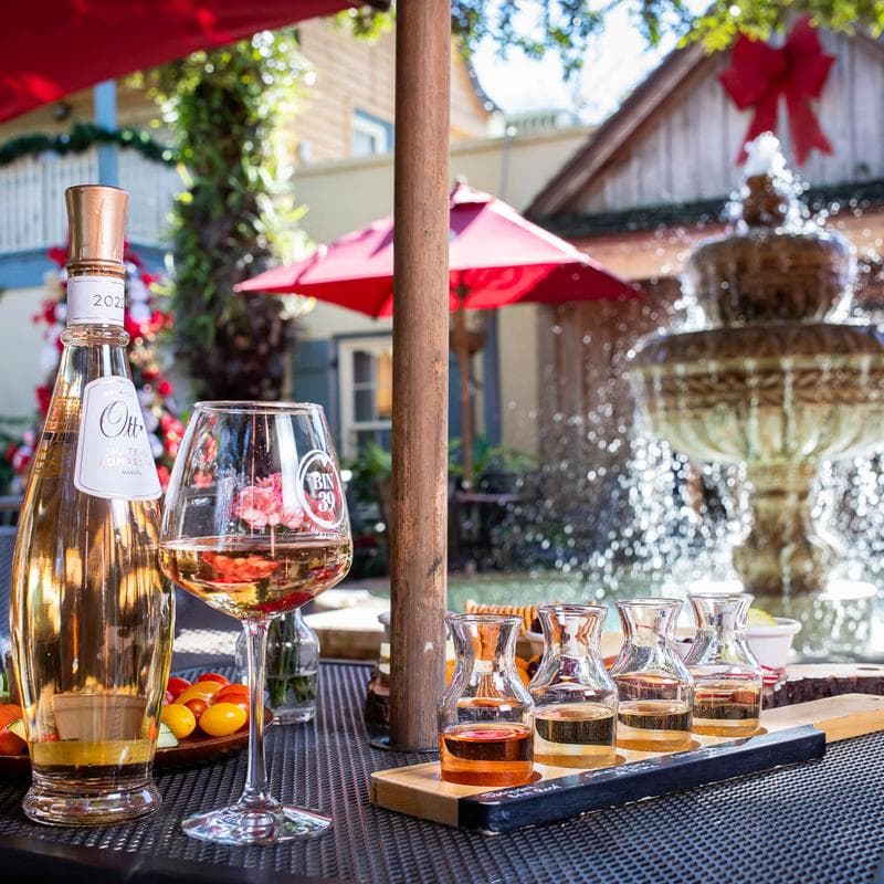 A bottle of rosé wine, a glass filled with wine, and tasting carafes arranged on a table near a fountain, with red umbrellas in the background.