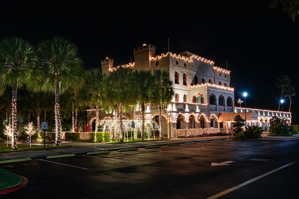 A historic building illuminated with festive lights at night, surrounded by palm trees.