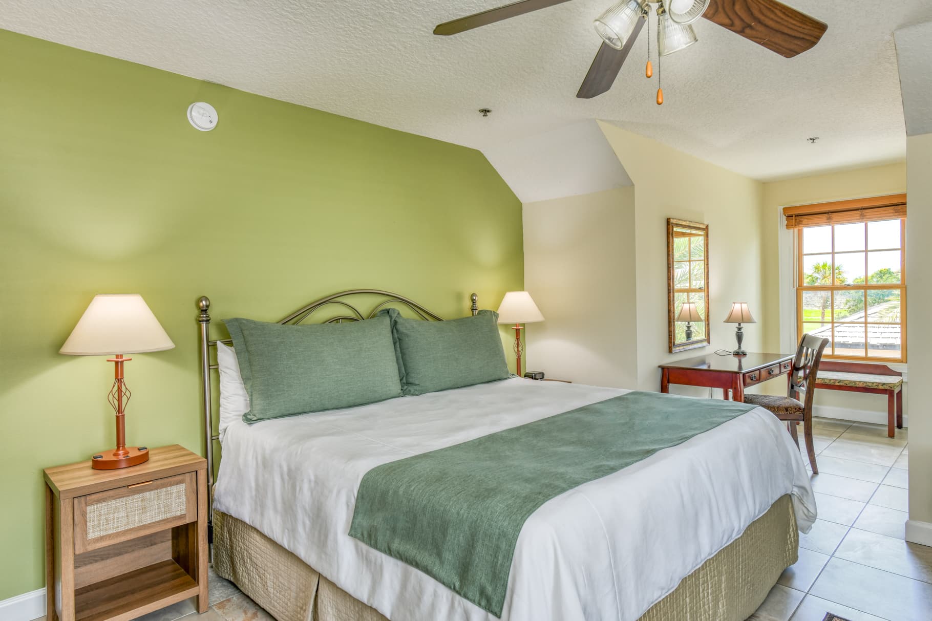 A cozy bedroom featuring a double bed, green walls, a desk, and windows with natural light.