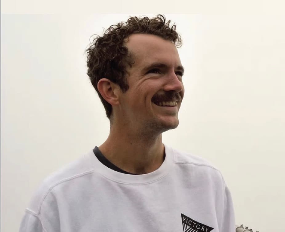 A smiling man with curly hair and a mustache wears a white sweatshirt against a foggy background.