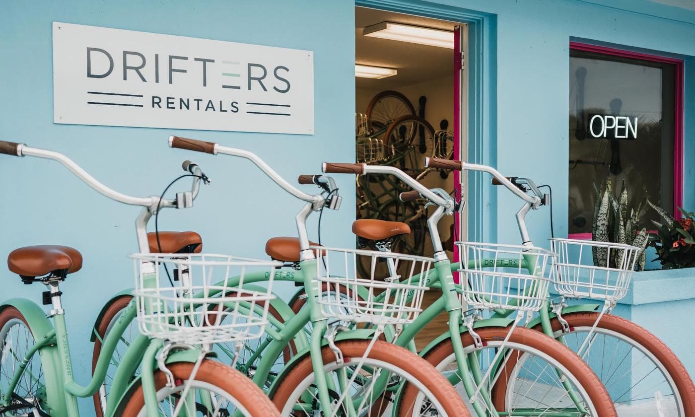 Four mint green bicycles with brown seats and white baskets are parked outside a rental shop with an "OPEN" sign.