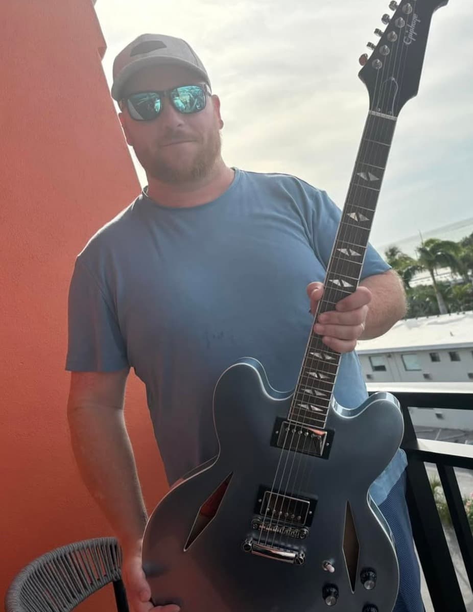 A man wearing a blue shirt and sunglasses holds a silver electric guitar on a balcony.