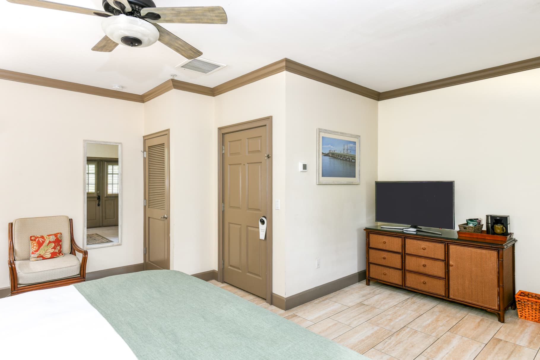 A light-colored bedroom featuring a bed, chair, TV cabinet with a television, and a mirror.