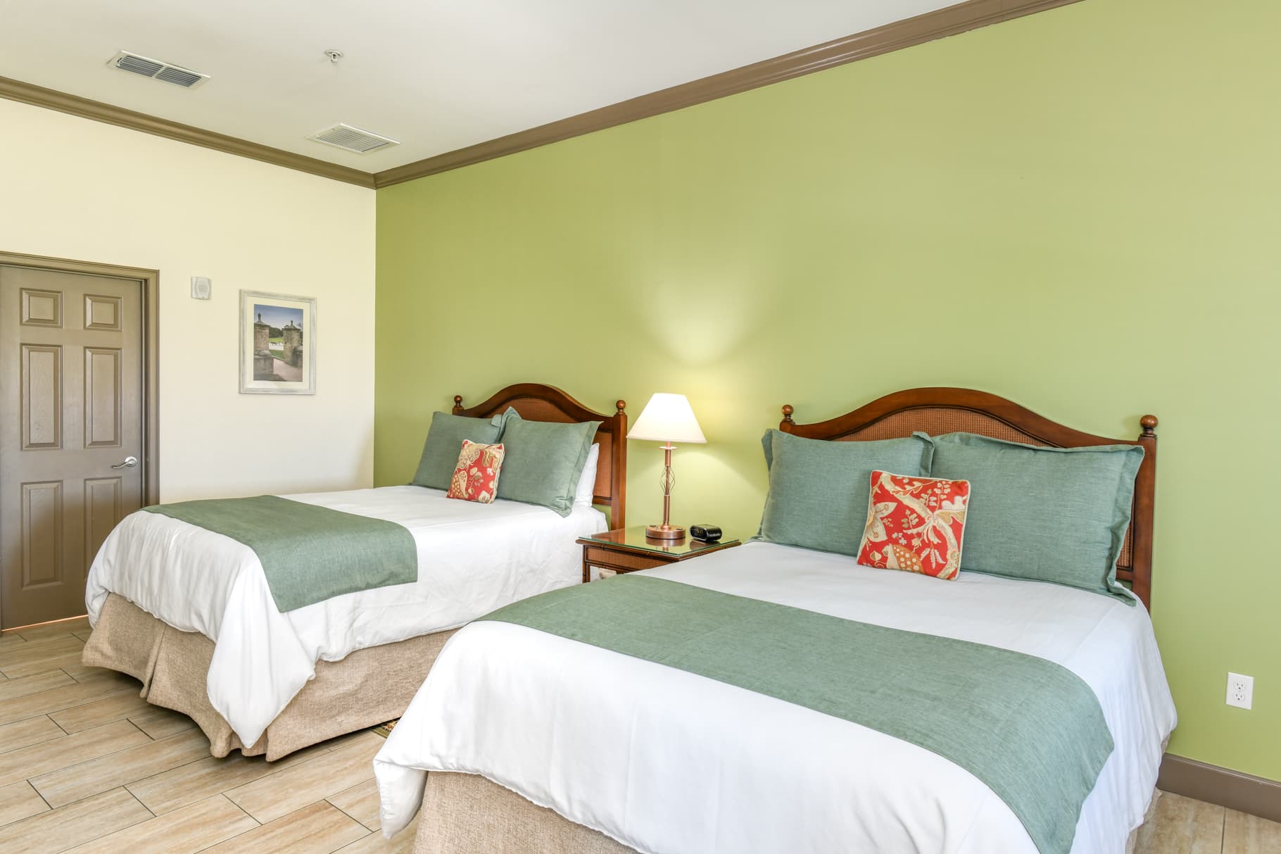 A bright hotel room featuring two beds with green accents and a lamp on a nightstand.