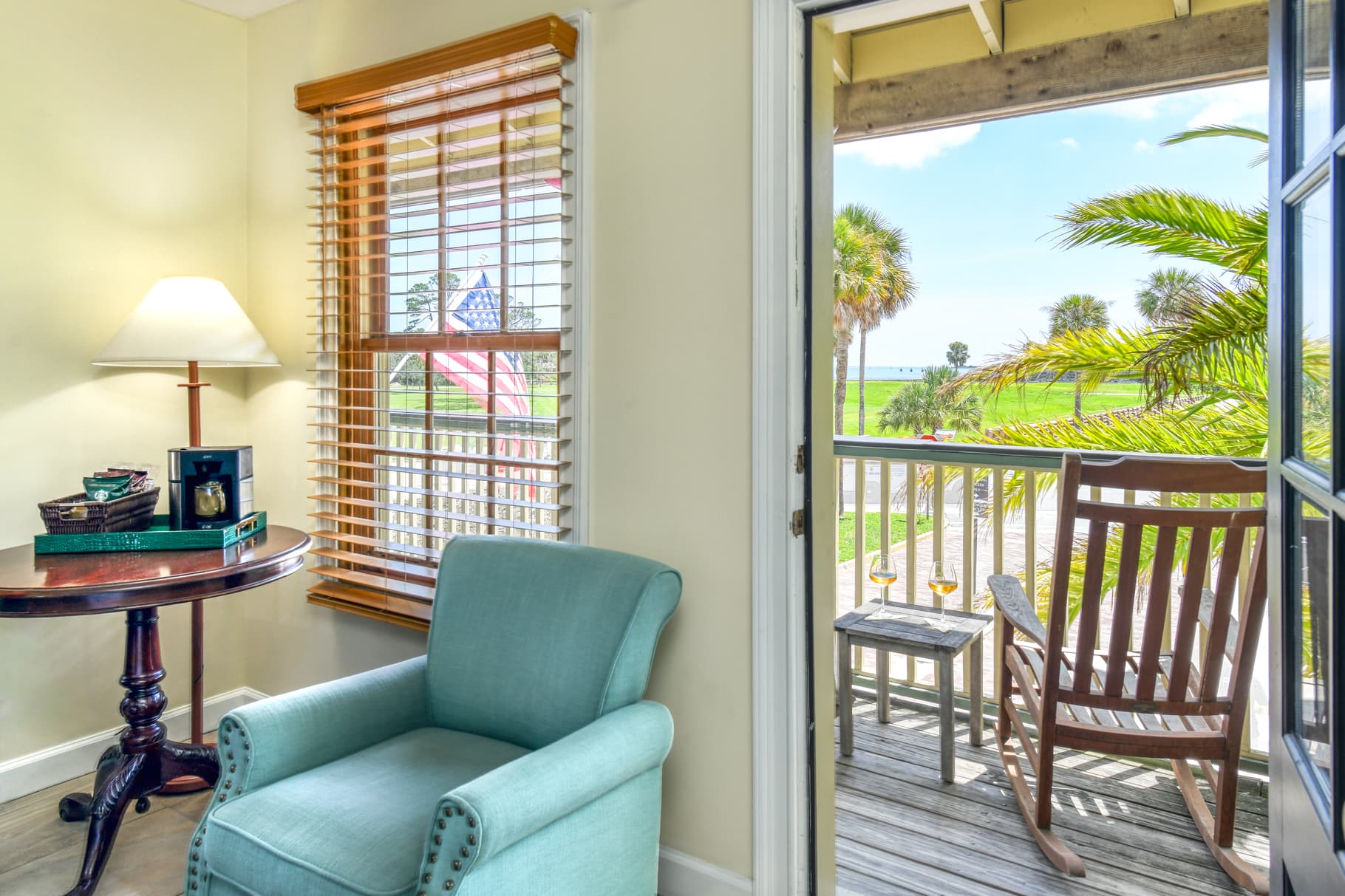 A cozy corner with a teal armchair, a side table, and a view of a sunny balcony with palm trees.