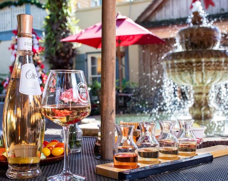 A bottle of rosé wine and a glass sit next to a flight of wine samples, with a fountain and patio umbrellas in the background.
