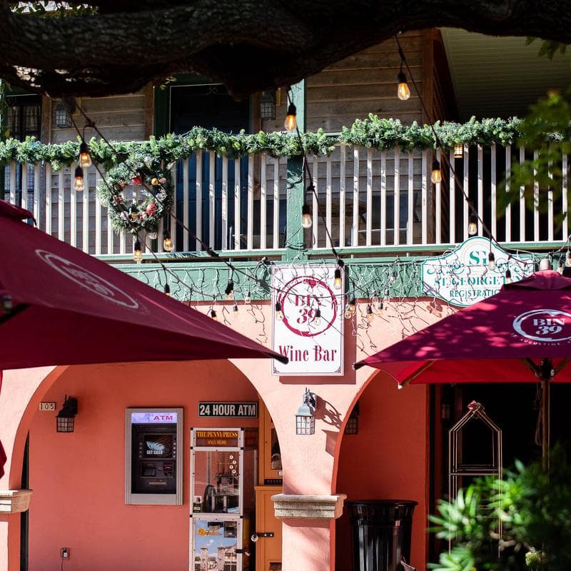 A wine bar with decorative greenery, outdoor seating, and a nearby ATM in a vibrant setting.