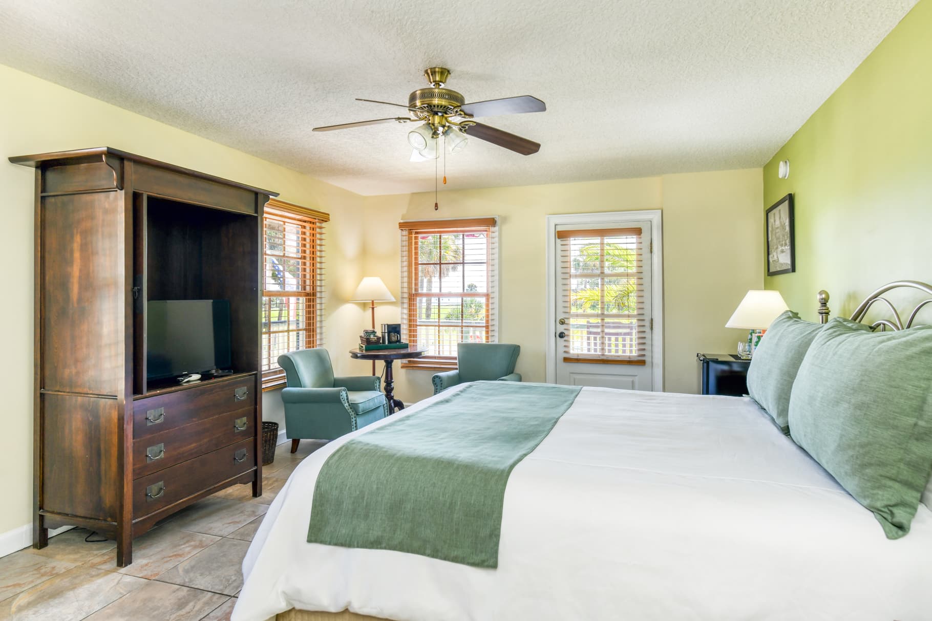 Brightly colored bedroom featuring a king-sized bed, a wooden armoire with a TV, and two cozy armchairs by the window.