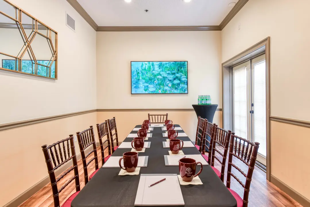 A conference room with a long black table set for a meeting, featuring chairs, mugs, and a large painting on the wall.