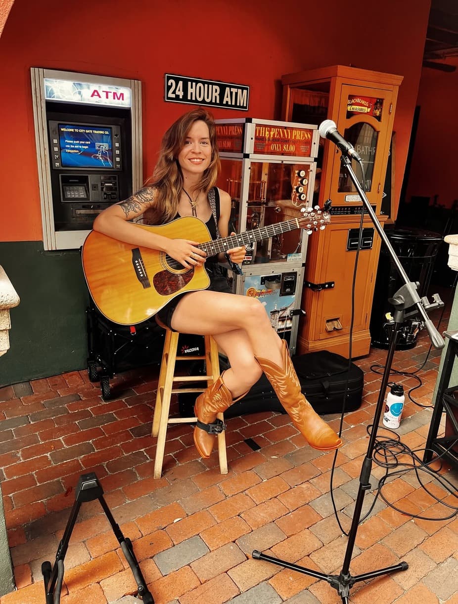 A woman sits on a stool playing an acoustic guitar in front of an ATM and other vintage machines.