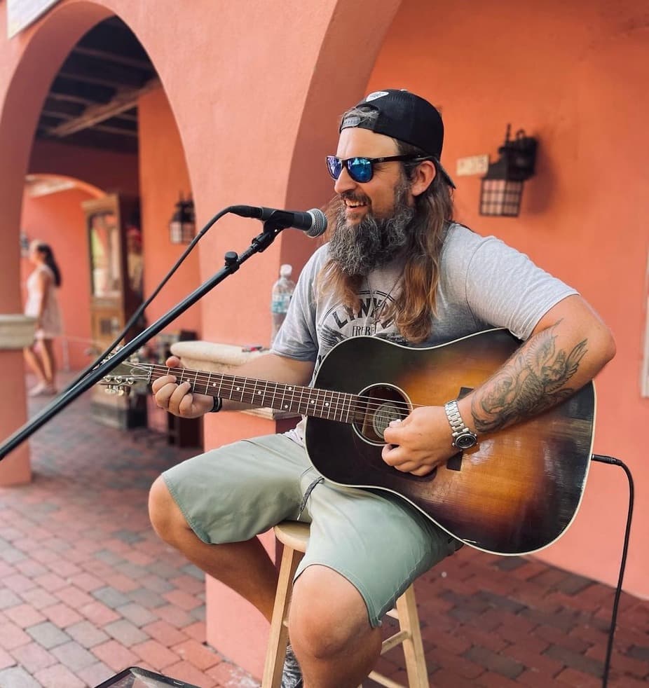 A musician with long hair and a beard plays guitar and sings on a stool against a warm, colorful background.