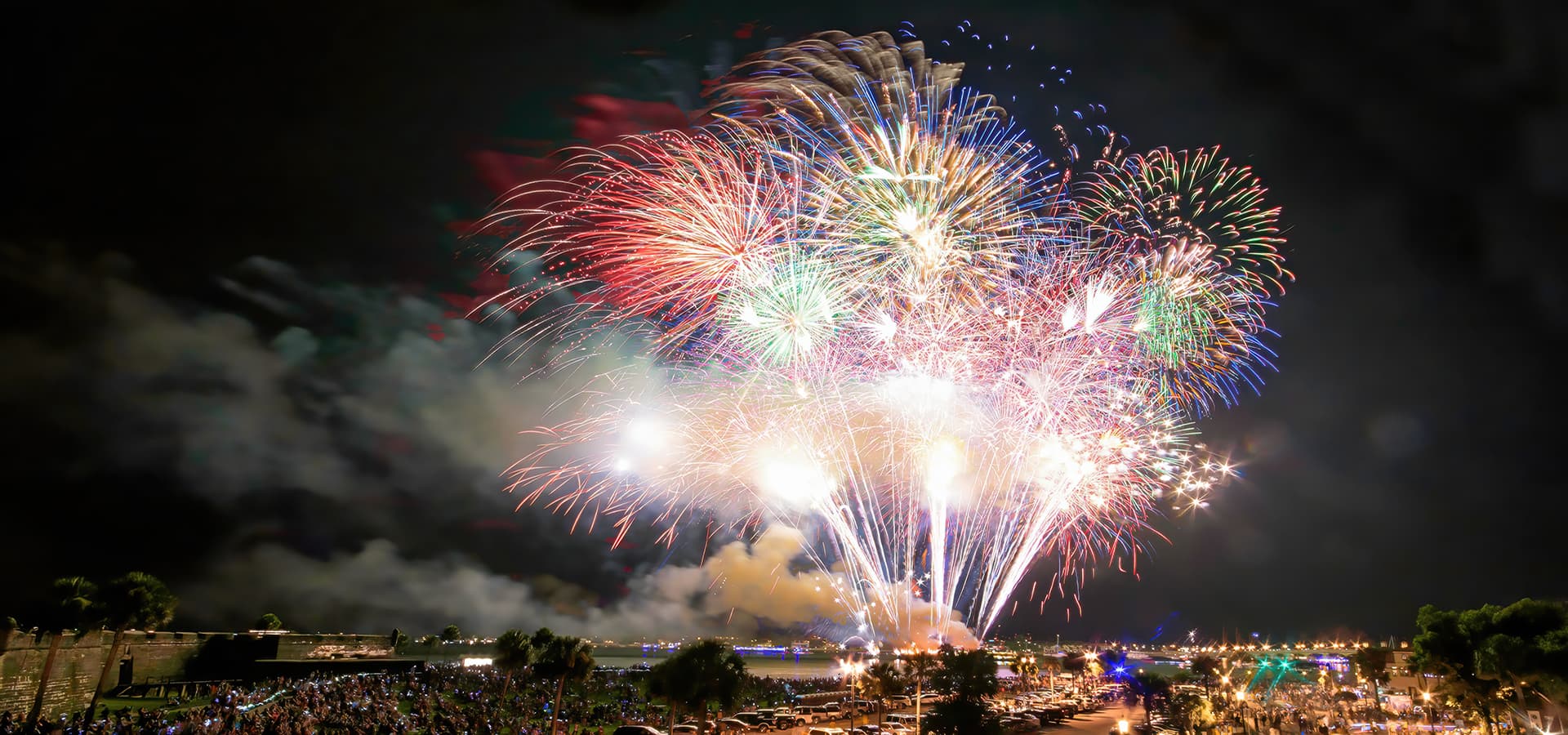 A vibrant display of colorful fireworks bursts in the night sky over a crowd near a waterfront.