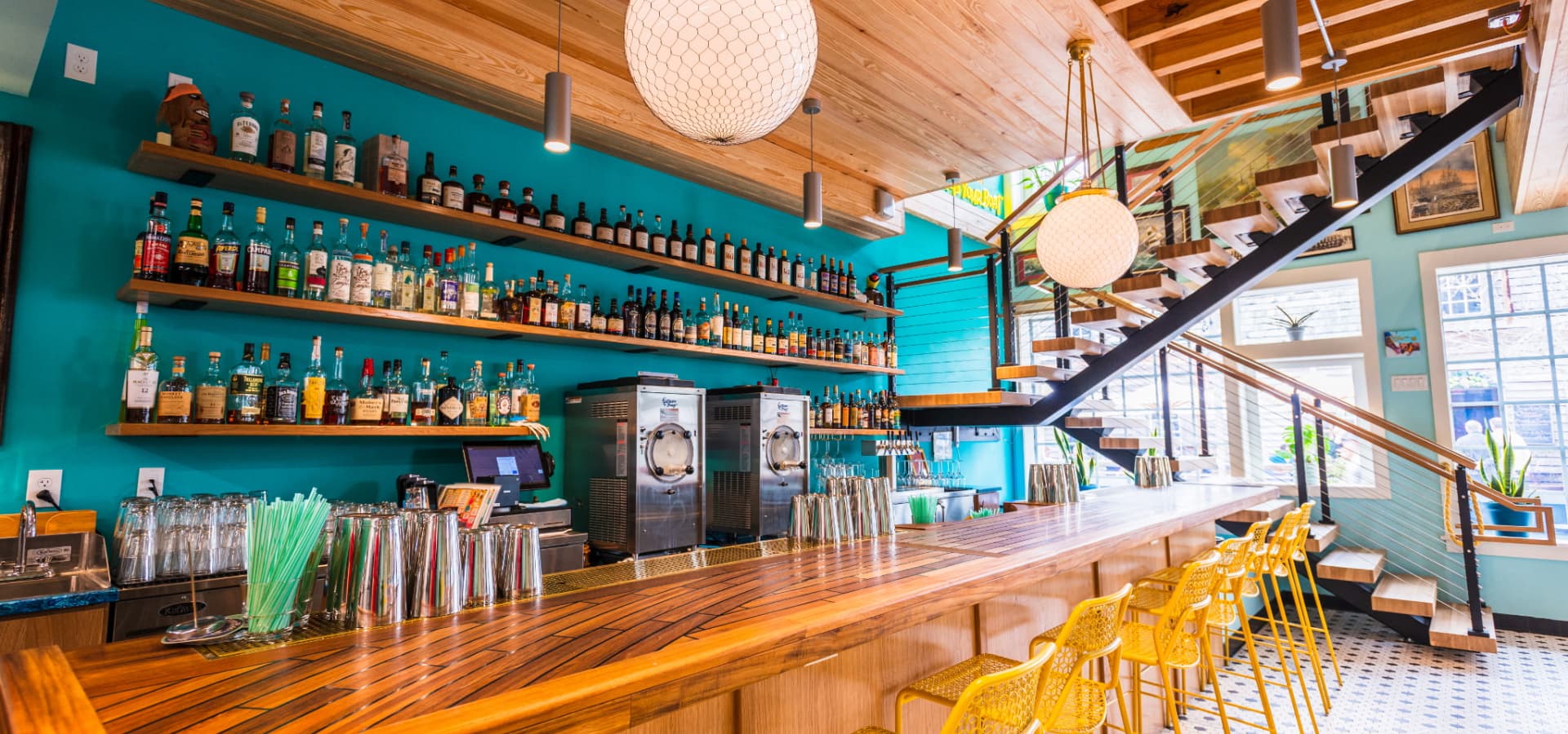 A brightly colored bar with a wooden countertop, numerous liquor bottles on shelves, and yellow stools.