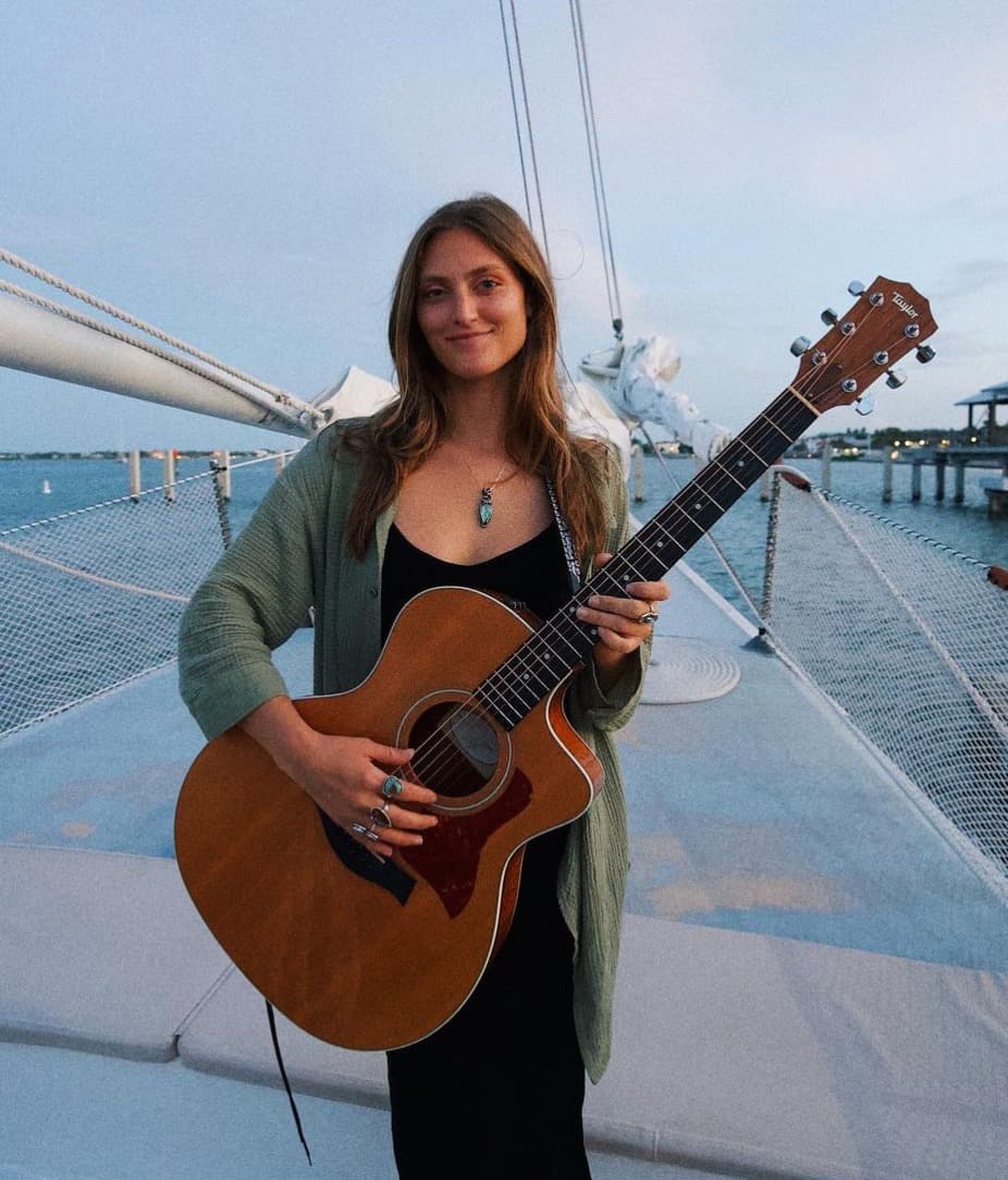 A woman smiles while holding an acoustic guitar on a boat, with the sea and sky in the background.