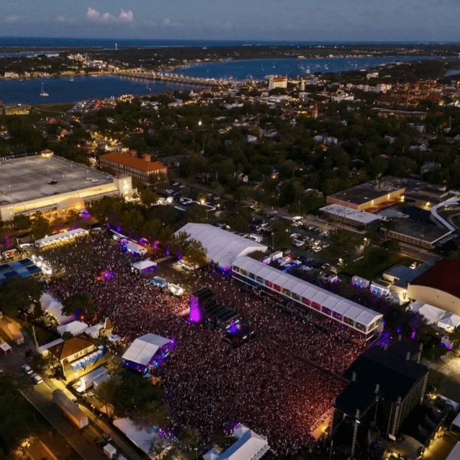 Aerial view of a large crowd at an outdoor festival, surrounded by tents and a waterfront cityscape at dusk.