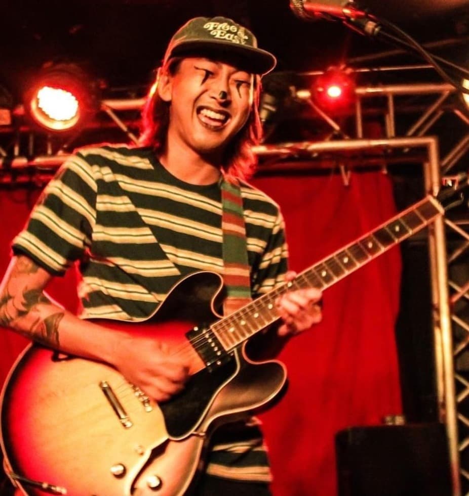 A smiling musician plays an electric guitar on stage under red lights.