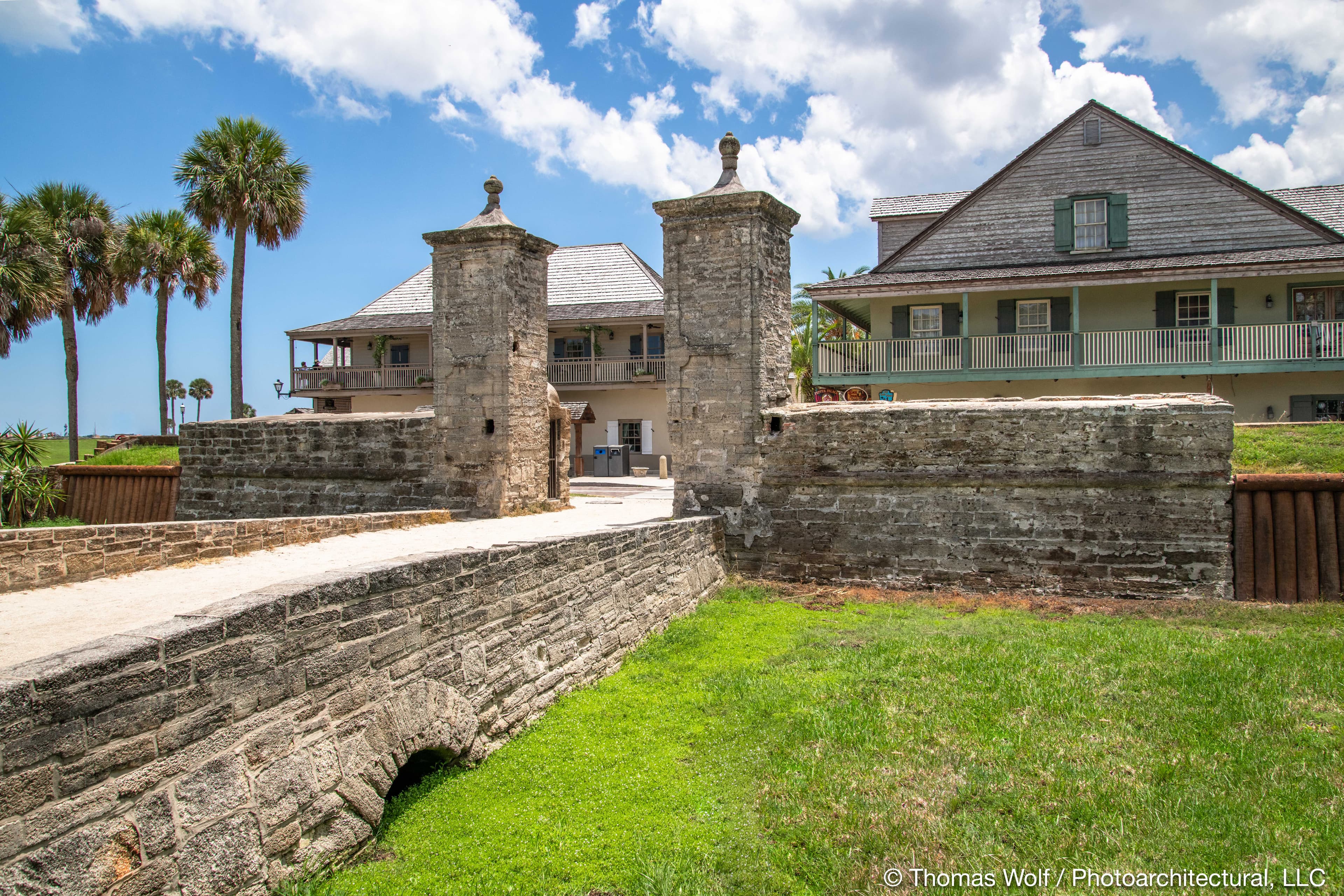 Historic stone entrance with two towers leading to a grassy area and a building in the background.