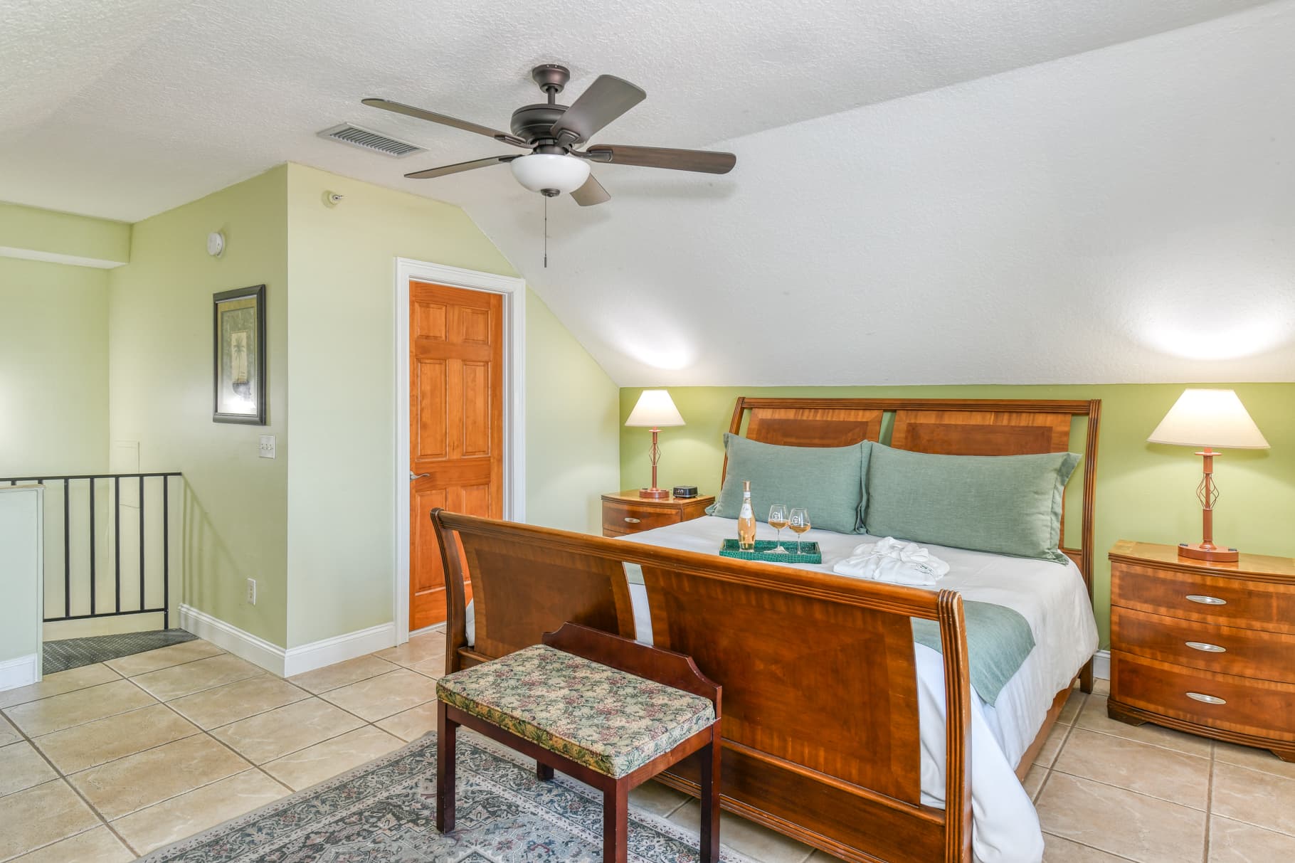 A cozy bedroom featuring a wooden bed, green walls, and a ceiling fan, with a small seating area and soft lighting.