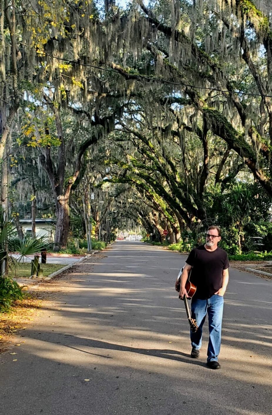A man walks down a tree-lined street carrying a guitar.