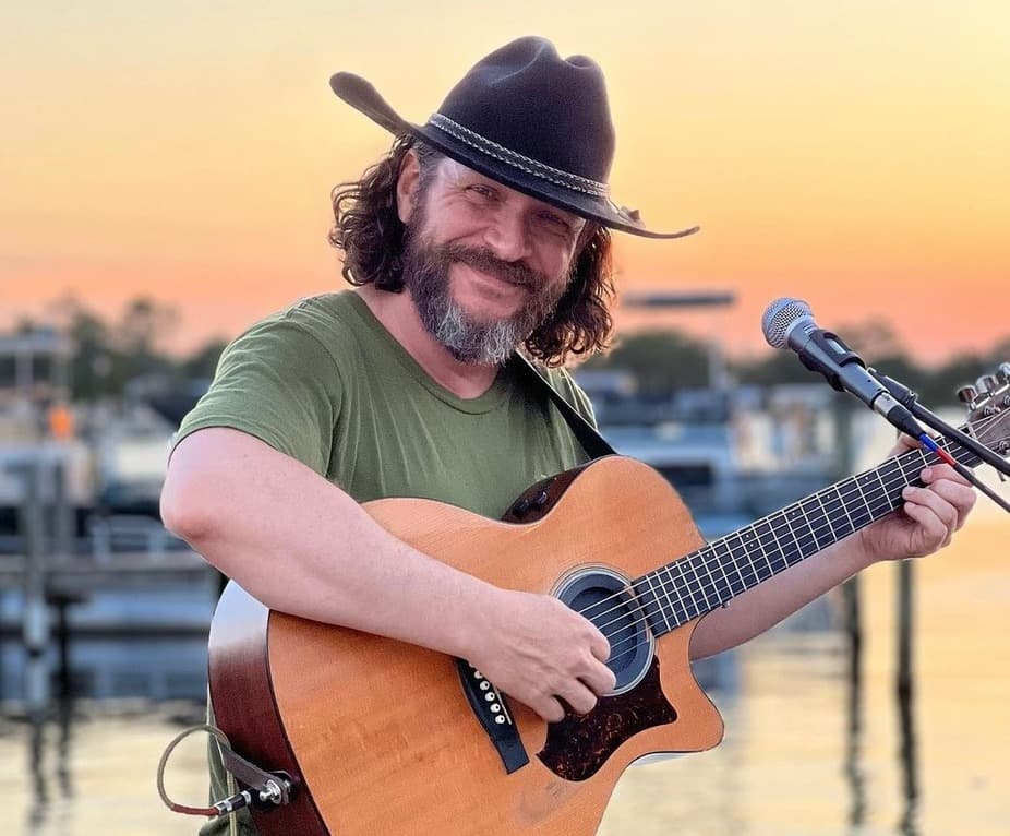 A man in a cowboy hat smiles while playing an acoustic guitar against a sunset backdrop.