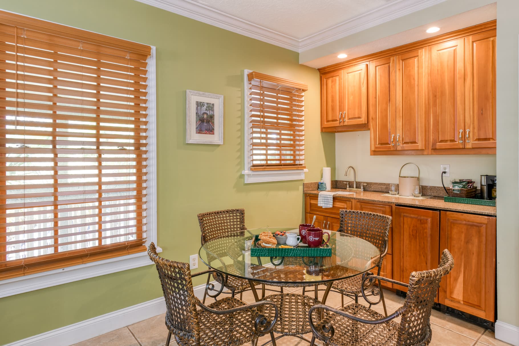 A cozy kitchen nook featuring a glass table surrounded by wicker chairs, with wooden cabinets and a green wall.