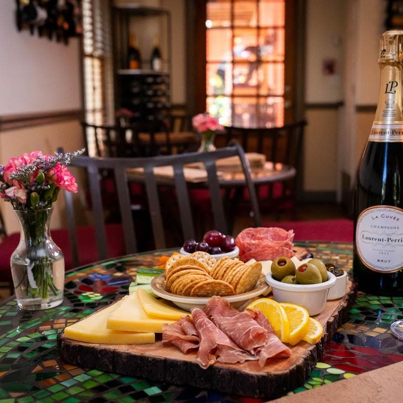 A charcuterie board with cheeses, meats, olives, crackers, and grapes, accompanied by a bottle of champagne, sits on a colorful table in a cozy restaurant setting.