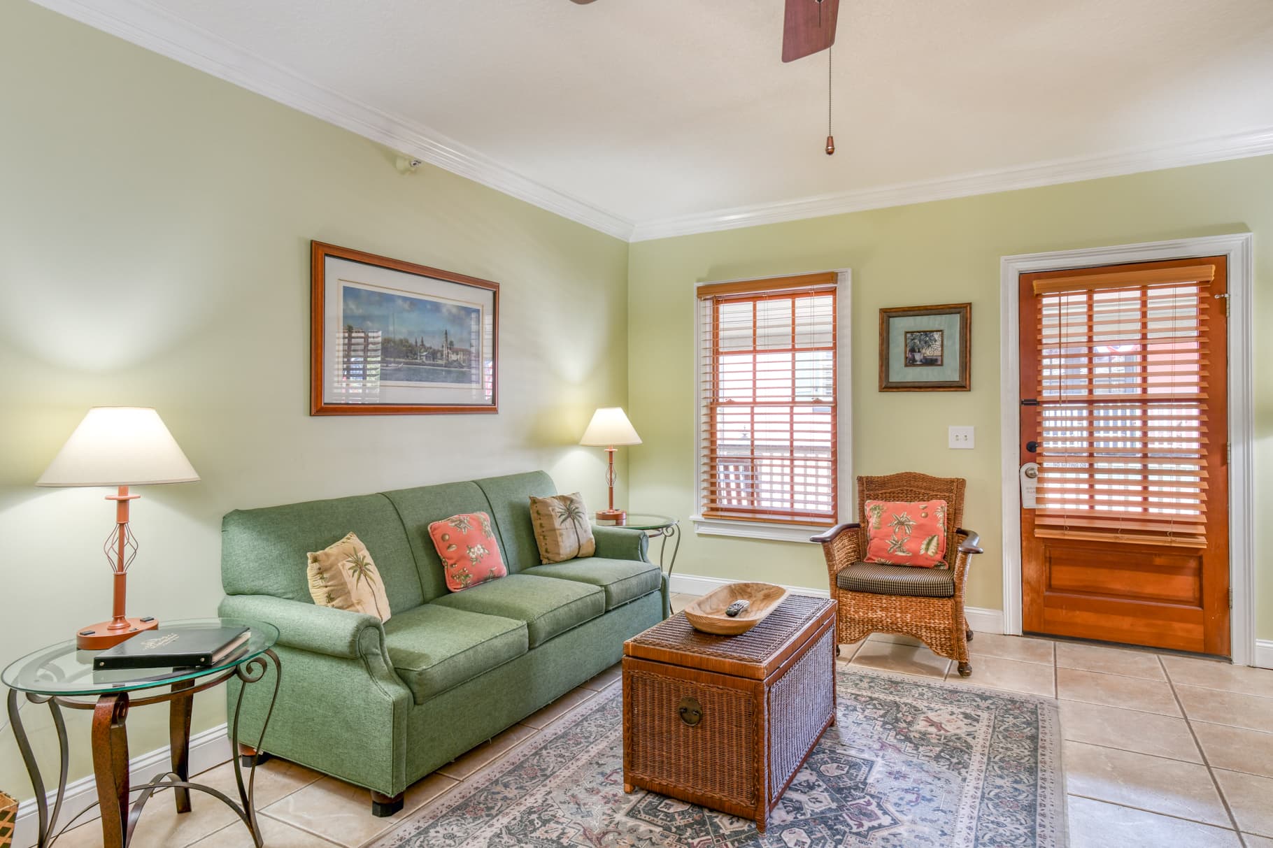 A cozy living room featuring a green sofa, wicker chair, decorative lamp, and a coffee table on tiled flooring.