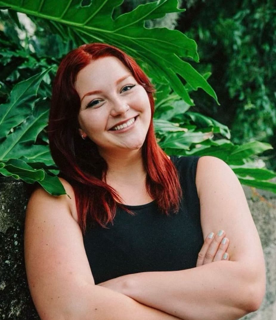 A smiling woman with long red hair poses confidently in front of large green leaves.