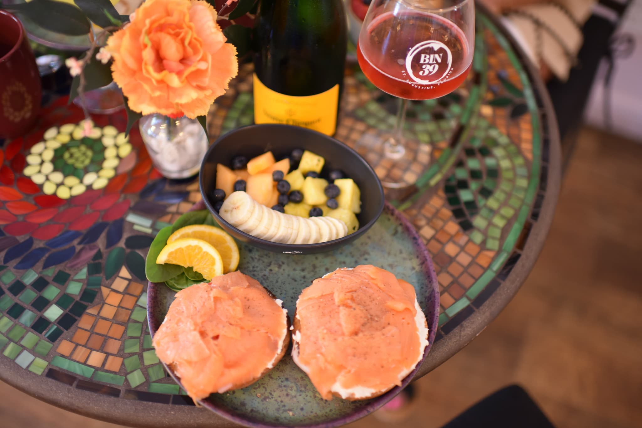 A colorful table setting featuring smoked salmon bagels, a fruit bowl, a glass of rosé, and a bouquet of flowers.