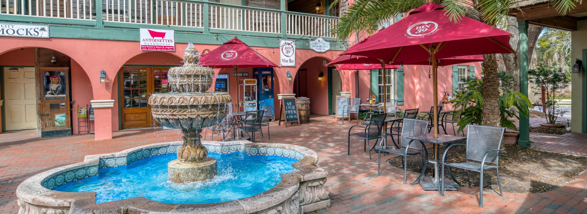 A stone fountain with blue water sits in a brick courtyard surrounded by cafes and shops under red umbrellas.