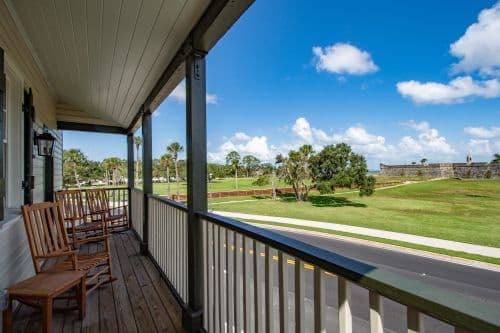 A porch with wooden rocking chairs overlooks a grassy area and blue sky with scattered clouds.