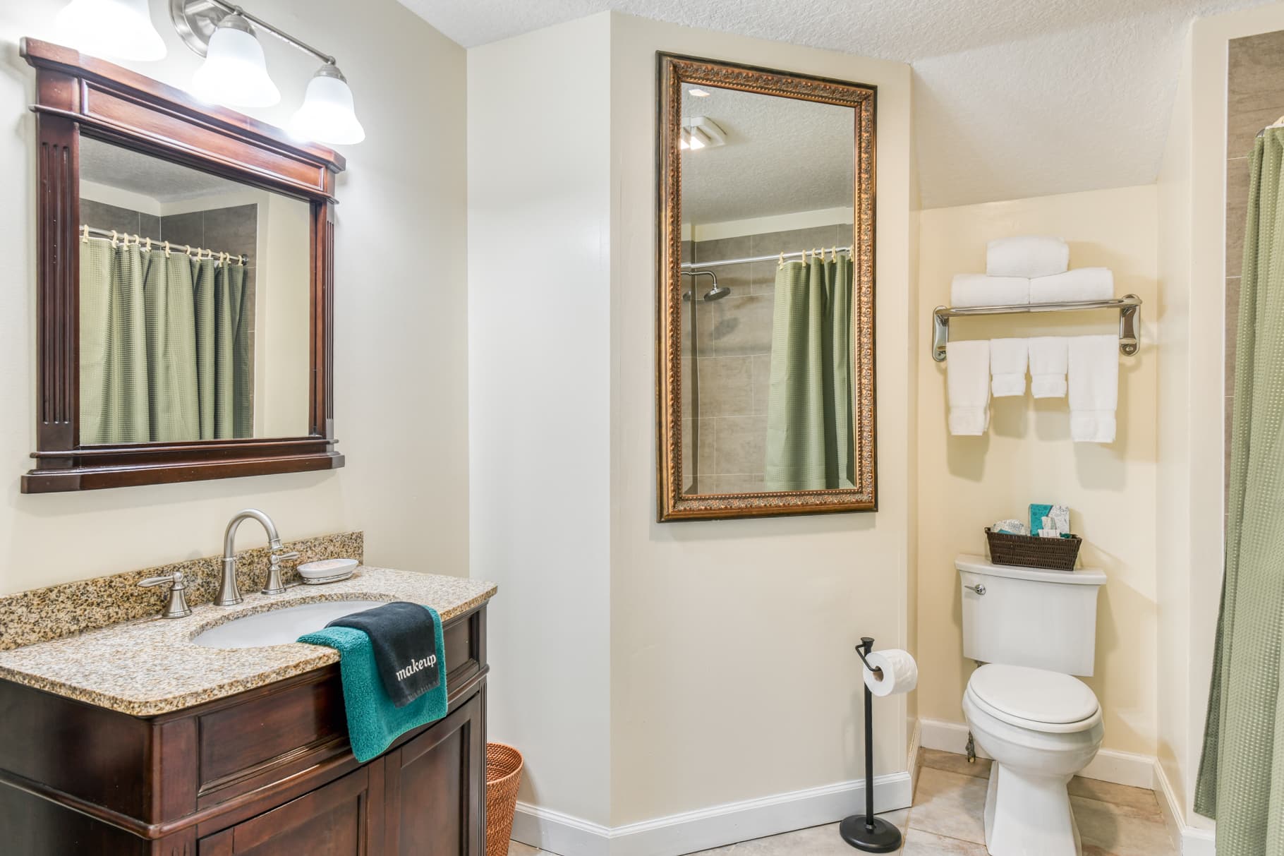 A well-lit bathroom featuring a dark wood vanity, a large mirror, green shower curtains, and neatly rolled white towels.