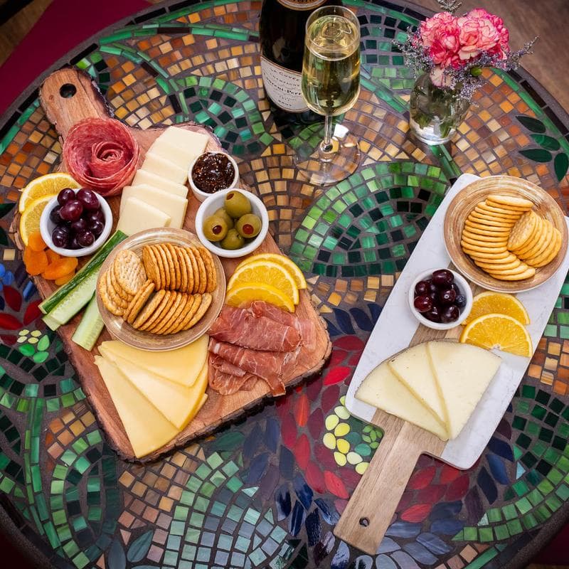 A mosaic-patterned table displaying two cheese and charcuterie boards, complemented by a glass of champagne and a small vase of flowers.