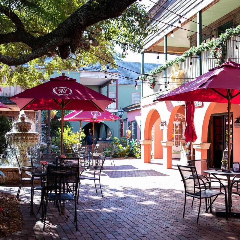A tranquil outdoor dining area with red umbrellas, a fountain, and colorful buildings.