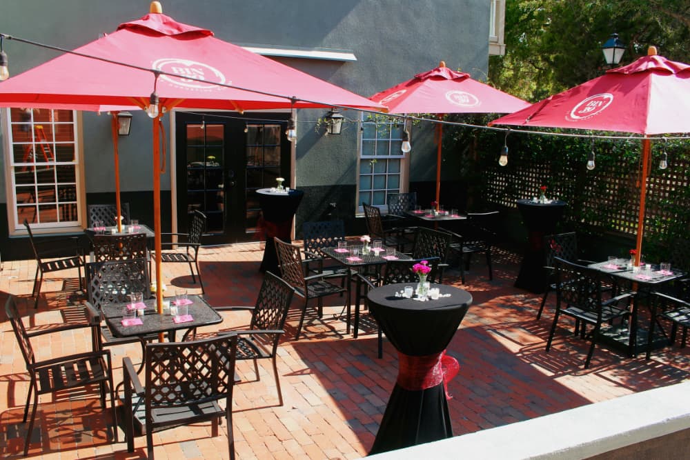 Outdoor dining area with black tables, vibrant red umbrellas, and decorative flowers on a brick patio.