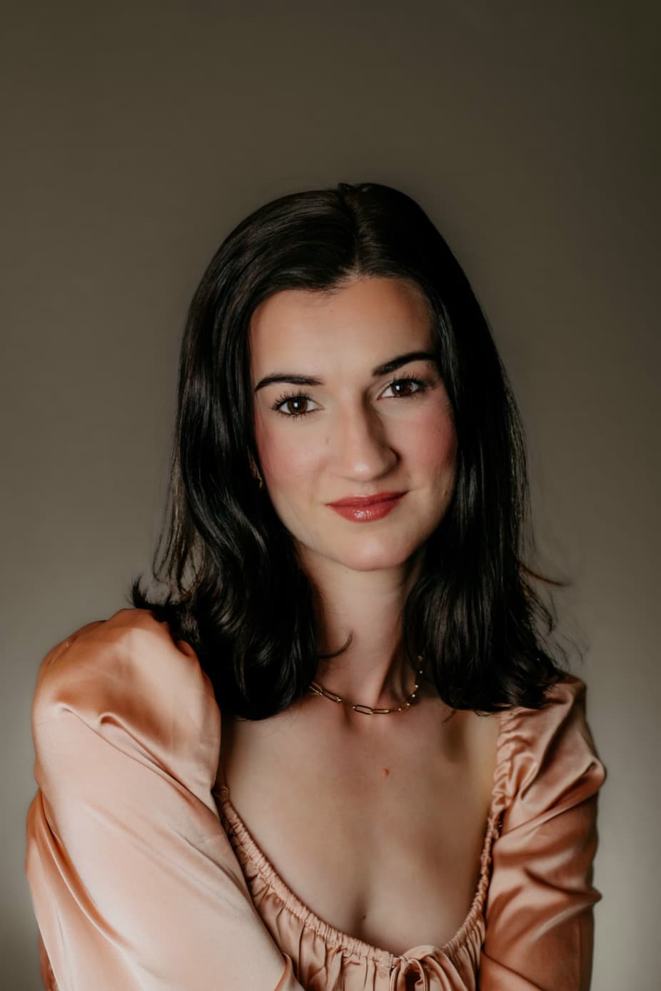 A woman with long dark hair smiles softly while wearing a silky peach blouse against a neutral background.