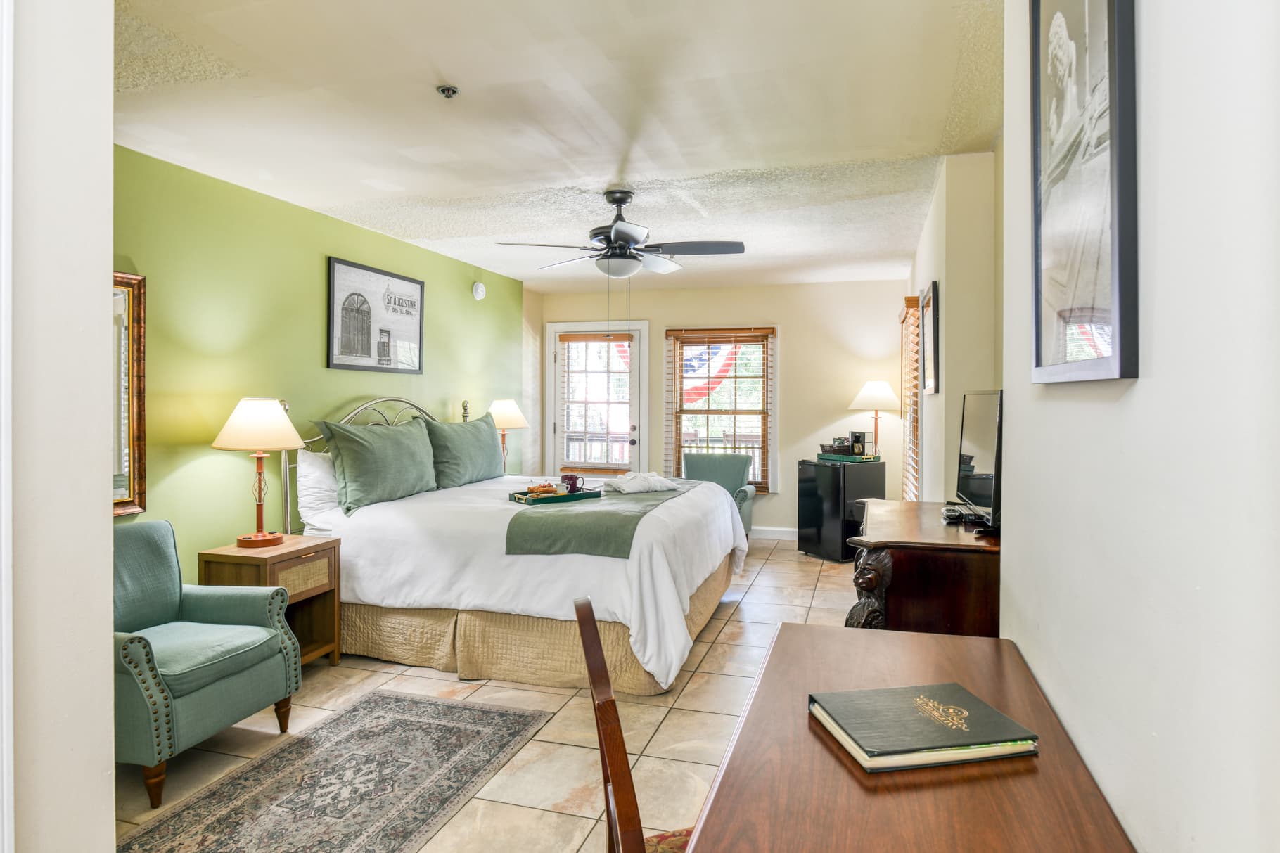 Cozy hotel room featuring a queen bed, decorative green walls, and a sitting area with a chair and desk.