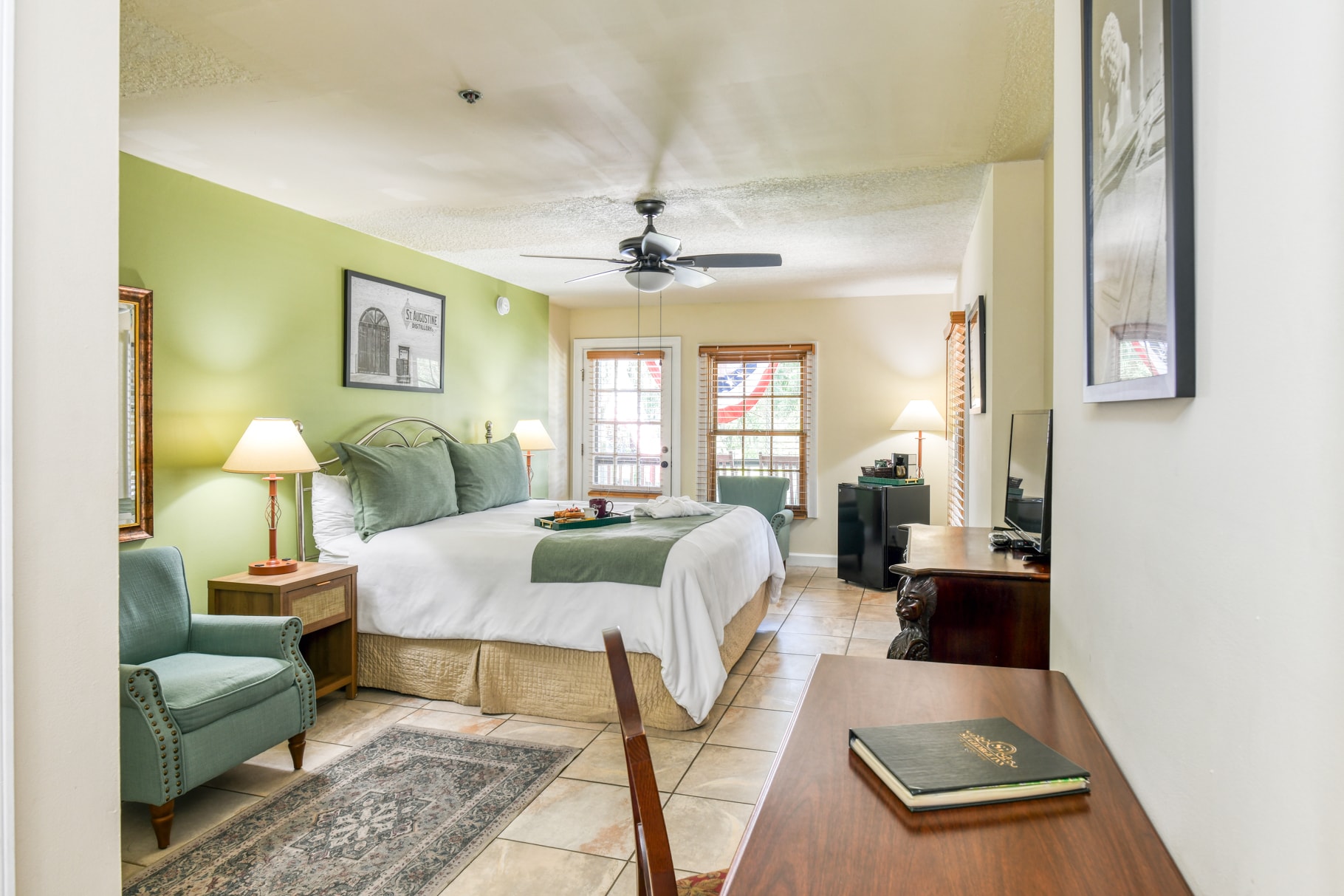 Cozy hotel room featuring a queen bed, decorative green walls, and a sitting area with a chair and desk.