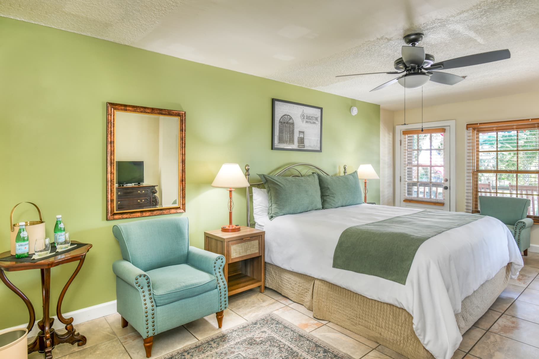 A cozy bedroom with a green accent wall, featuring a large bed, a mirror, a ceiling fan, and a seating area.