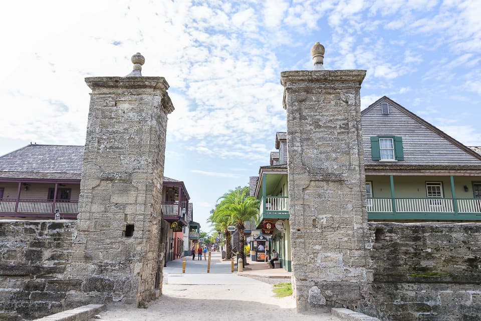 Historic stone entrance flanked by buildings and palm trees under a partly cloudy sky.